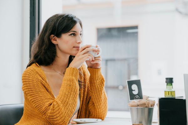 Valley Creek Woman in a mustard sweater holding a cup, sitting by a window in a café.